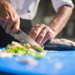 Chef fatia um peixe fresco sobre uma tábua azul, em uma cozinha profissional, durante o preparo culinário.