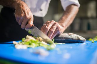 Chef fatia um peixe fresco sobre uma tábua azul, em uma cozinha profissional, durante o preparo culinário.