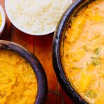 Moqueca brasileira colorida servida em panela de barro, acompanhada de outros pratos sobre uma mesa de madeira