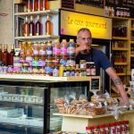 Comerciante diante de uma banca de feira com diversos produtos gourmet expostos.